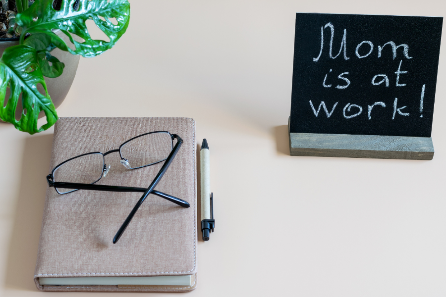 Planner on a desk with eyeglasses on top next to a sign that says, Mom is at work! Mom has learned how to start a mom business. 